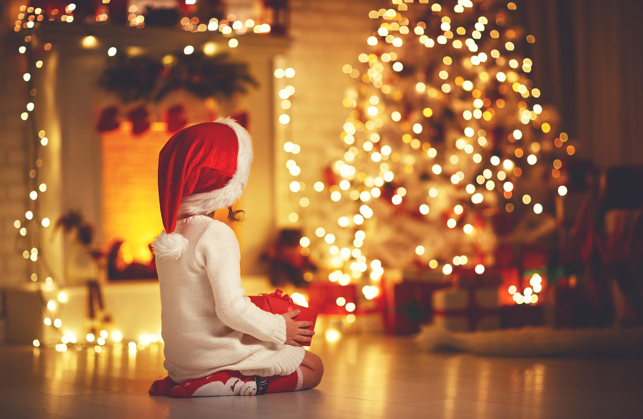 Bambina seduta con un regalo tra le mani che guarda l'albero di natale